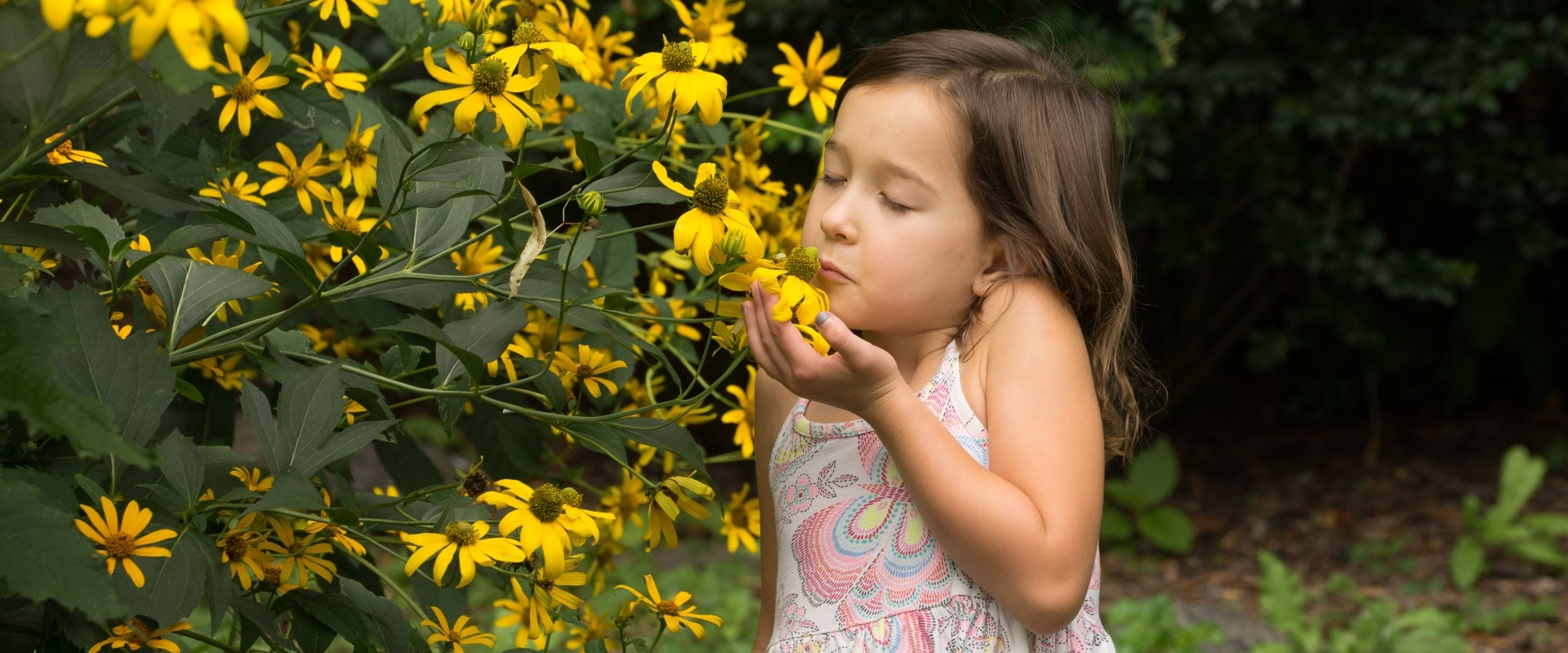 A young girl holds a yellow flower in her hand and inhales with her eyes closed.