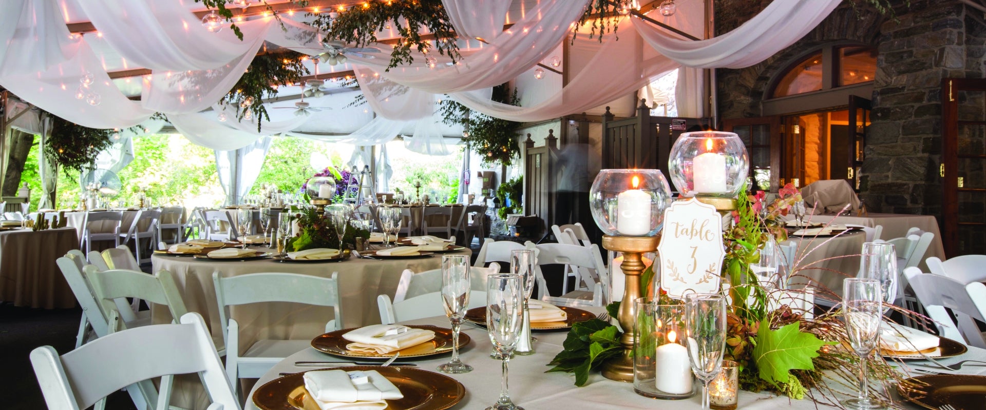 An outdoor tent decorated with lights, linens, candles, and table settings for a wedding.