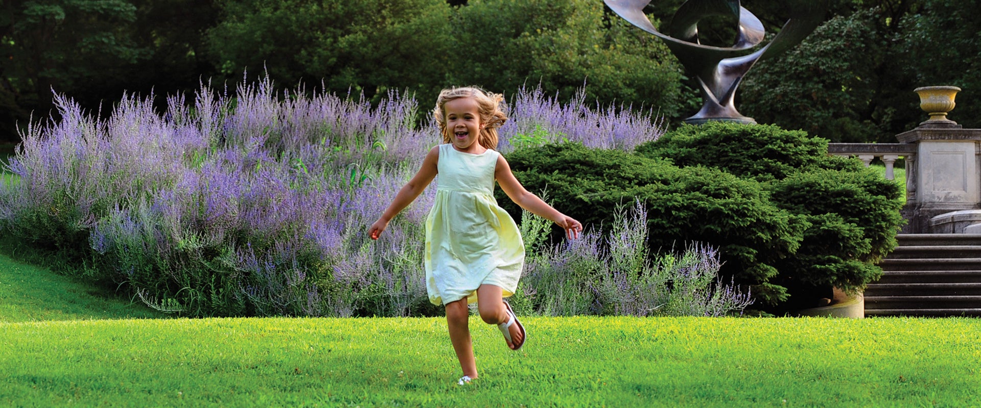 A young girl in a light yellow dress runs smiling through a green space in a public garden.