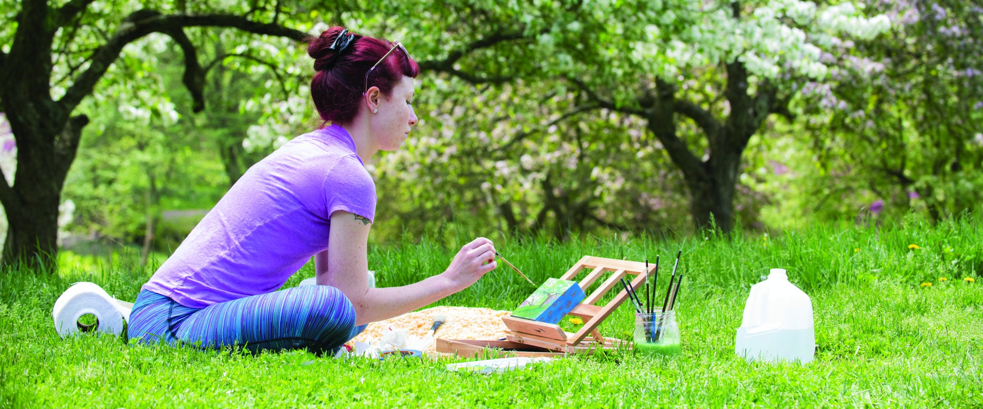 A woman sits outside in the grass surrounded by blooming trees painted on a small canvas. 