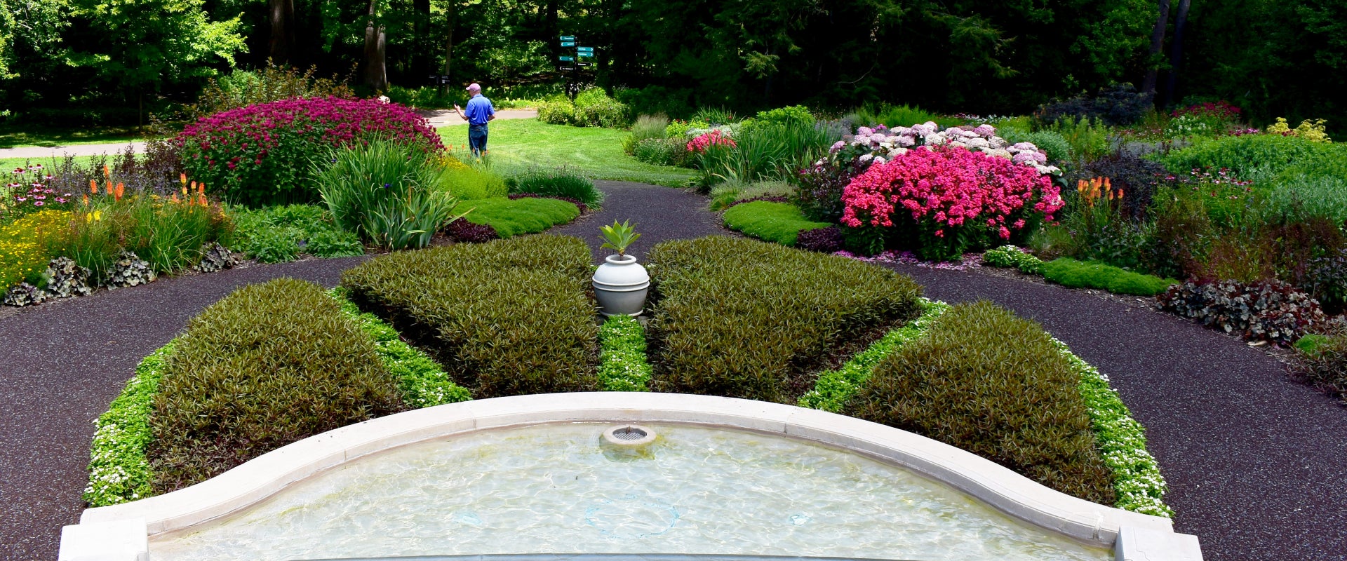 A garden designed with multiple beds filled with colorful flowers and a fountain with a man in the background. 
