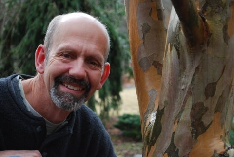 A man with a short beard smiles next to a tree trunk.
