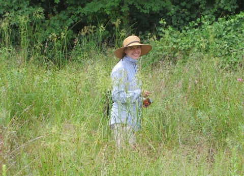 A woman in a wide-brimmed hat stands in a meadow smiling. 