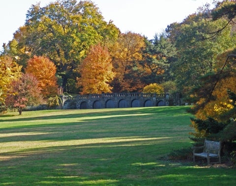 An expansive lawn with fall foliage trees and a long stone bridge.