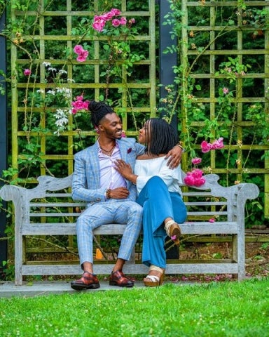 An engagement photo of a couple sitting on a bench, holding each other, with roses in the background.