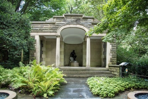 A traditional classically-styled stone garden building with a terrace and statue, surrounded by green plants and trees.