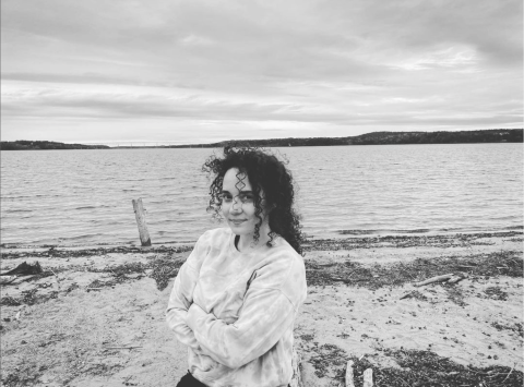 A black-and-white photo of a woman with dark, curly hair smiling and crossing her arms across her chest with water in the background.