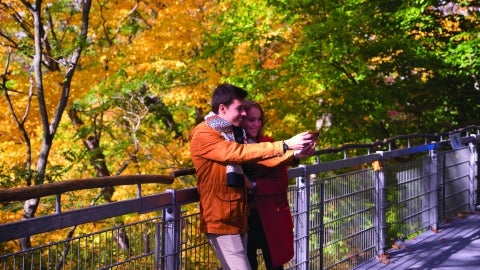 Two students hold up a phone to take a selfie in front of fall foliage. 