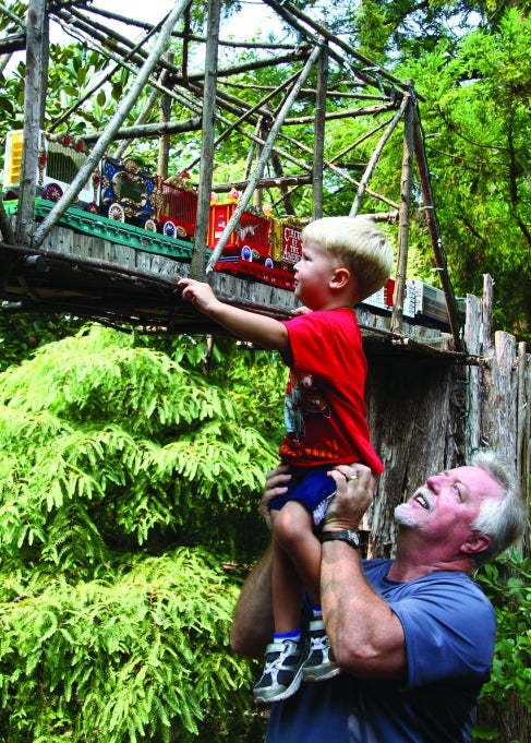 A man holds a young boy up toward a miniature elevated train bridge with a model train riding by. 