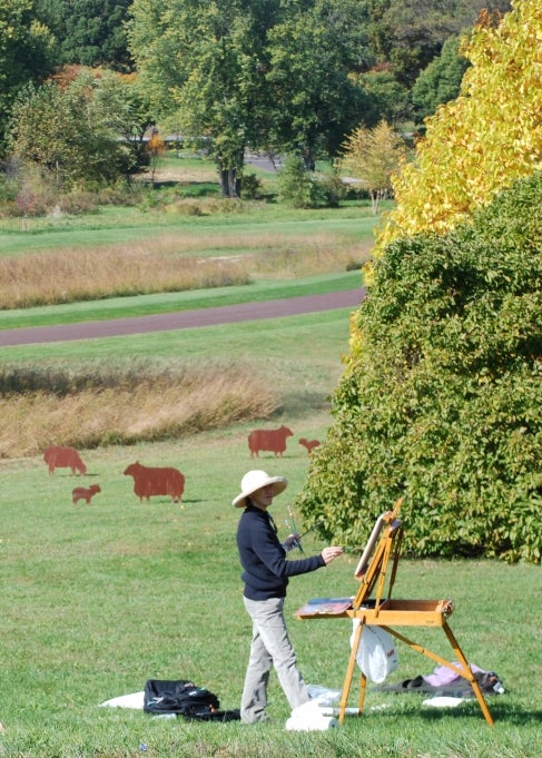 A woman stands in a public garden and paints on an easel. 