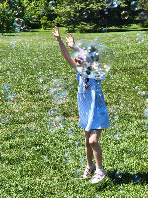 A young girl in a blue striped dress and sunglasses stands with her arms raised in a field of bubbles.