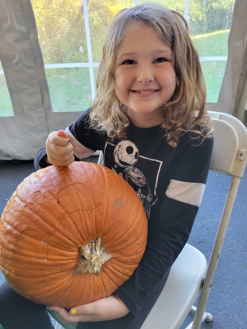 A young girl wearing a black and white Halloween shirt smiles while she carves a pumpkin. 