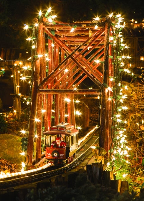 A miniature train display at night with Santa riding on a car through a trestle decorated with lights. 