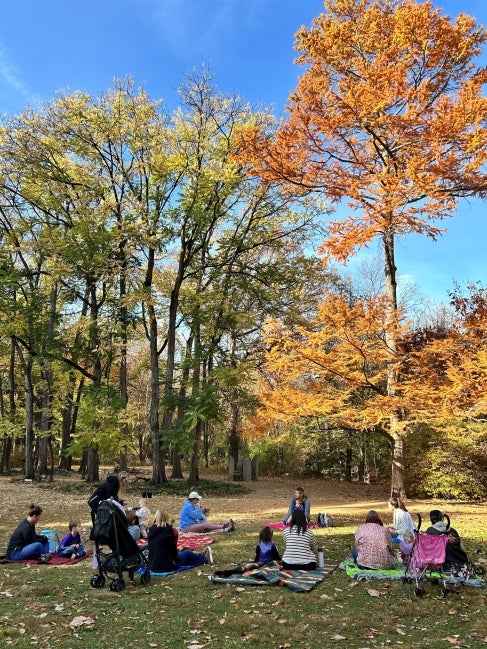A group of families sit on blankets the ground outdoors in fall for a class. 