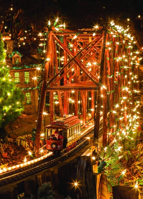 A miniature train display of a wooden bridge decorated in lights with a Christmas train car riding through with Santa in it.