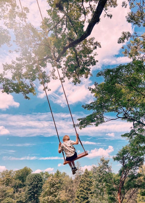 The back of a child on a swing surrounded by trees on a sunny day.