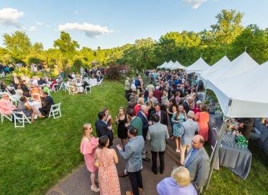 A large group of people mingle with drinks and appetizers in a public garden, at tables and under white tents. 