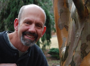 A man with a beard smiles next to a tree trunk. 