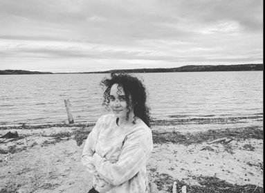A black and white photo of a woman with dark curly hair standing in front of a body of water. 