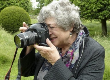 A woman with white hair holds a camera up to her face with greenery in the background. 