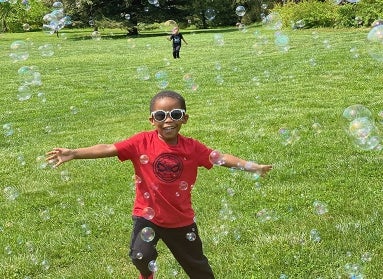 A young boy in sunglasses runs with his arms wide through bubbles in a large, green field. 