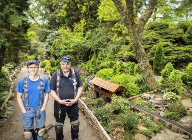 A young man and an older man wearing thick suspended and hats stand outdoors within a garden railway featuring miniature trains and structures. 