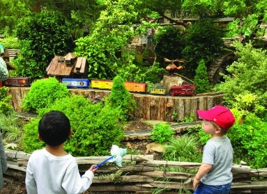 Two young boys, one holding a pinwheel, watching a miniature train rides by in a garden railway display.
