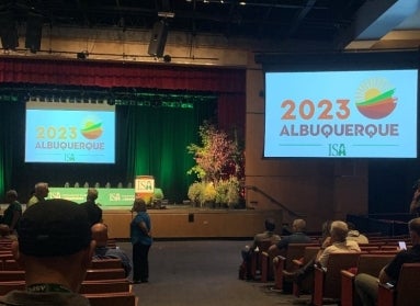 A dimly lit auditorium with people sitting and standing and a projected that reads, "2023 Albuquerque ISA."