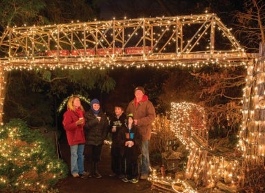 A family looking up at a miniature trestle decorated with lights as a model train rides through.
