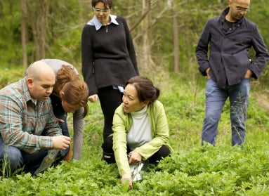 A woman kneels on the ground outdoors, with four others surrounding her, pointing toward a plant on the ground and smiling. 
