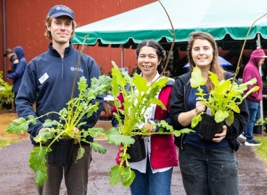 Three people hold plants outside of a barn while smiling. 
