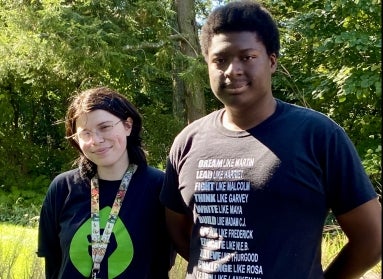 Two young students stand next to each smiling at the camera in a green outdoor setting.