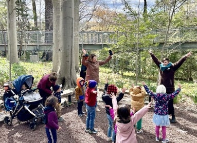 A photo of a group of children and a few adults outdoors in coats with their arms raised and smiling. 