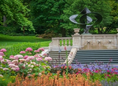 A fountain of lions heads spewing water with a large geometric sculpture in the background and flowers in the foreground. 