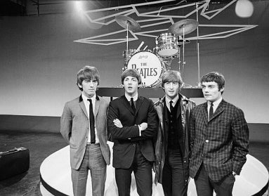 The musical group The Beatles standing together on a stage with a drunk in the background and a kickdrum that reads, "The Beatles."