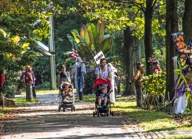 A woman pushes a stroller on a path surrounded by greenery and trees as well as scarecrows. 