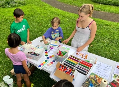 A group of four children stand around a table outdoors water coloring and drawing. 