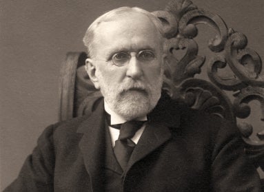 A black and white photograph of an older man with white hair and glasses dressed in a suit seated in an ornate wooden chair.