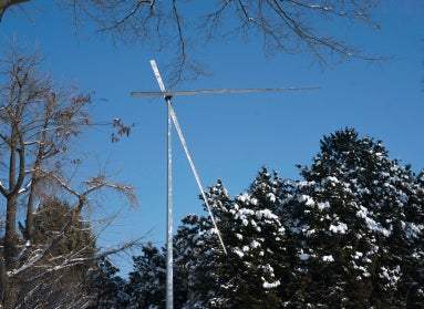 A tall, metal kinetic sculpture set against a backdrop of evergreen trees dusted in snow and blue sky.