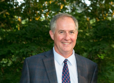 Man in a dark gray suit and blue and red tie stands in front of a tree with green leaves