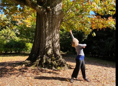 A woman dances outdoors under a large oak tree in fall with leaves covering the ground.