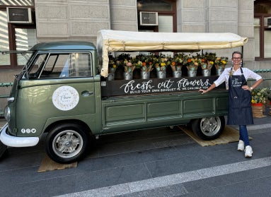 A woman wearing an apron leans again a small green truck with flowers for sale in metal buckets and a hand-painted sign that reads, "Fresh Cut Flowers." 