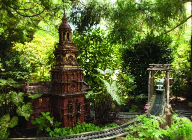 A miniature replica of Philadelphia's Independence Hall made of natural materials set  out doors next to a small bridge with a miniature train riding through. 