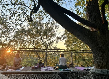 Three woman sit on yoga mats on an outdoor platform in the trees at dusk. 