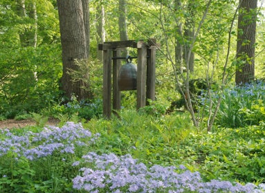 A large sculpture of a bell stands in a public garden surrounded by blue and white flowers in spring. 
