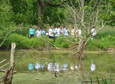 A group of students stand in an outdoor natural area with their reflection mirrored in a pond. 