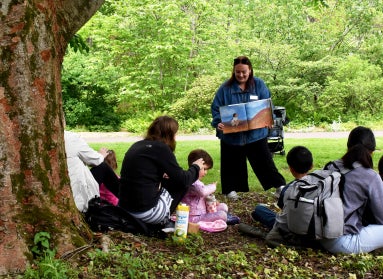 A young woman holds up a picture book to a small group of children and caretakers in an outdoor setting.