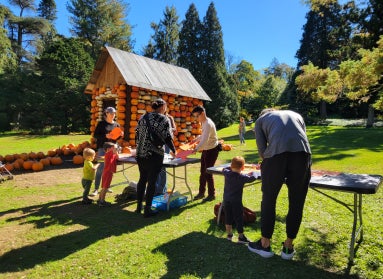 A group of people stand outside at a table doing crafts. In the background is a small cottage covered in pumpkins. 