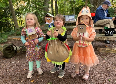 Three small children dressed in Halloween costume hold up crafts in an outdoor classroom. 