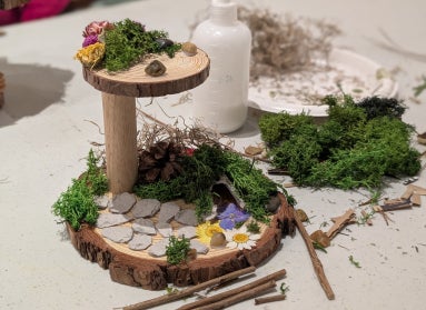 A small fairy house craft in mid-construction made out of natural materials including wood, moss, stones, and flowers, set on a white table with a bottle of glue. 